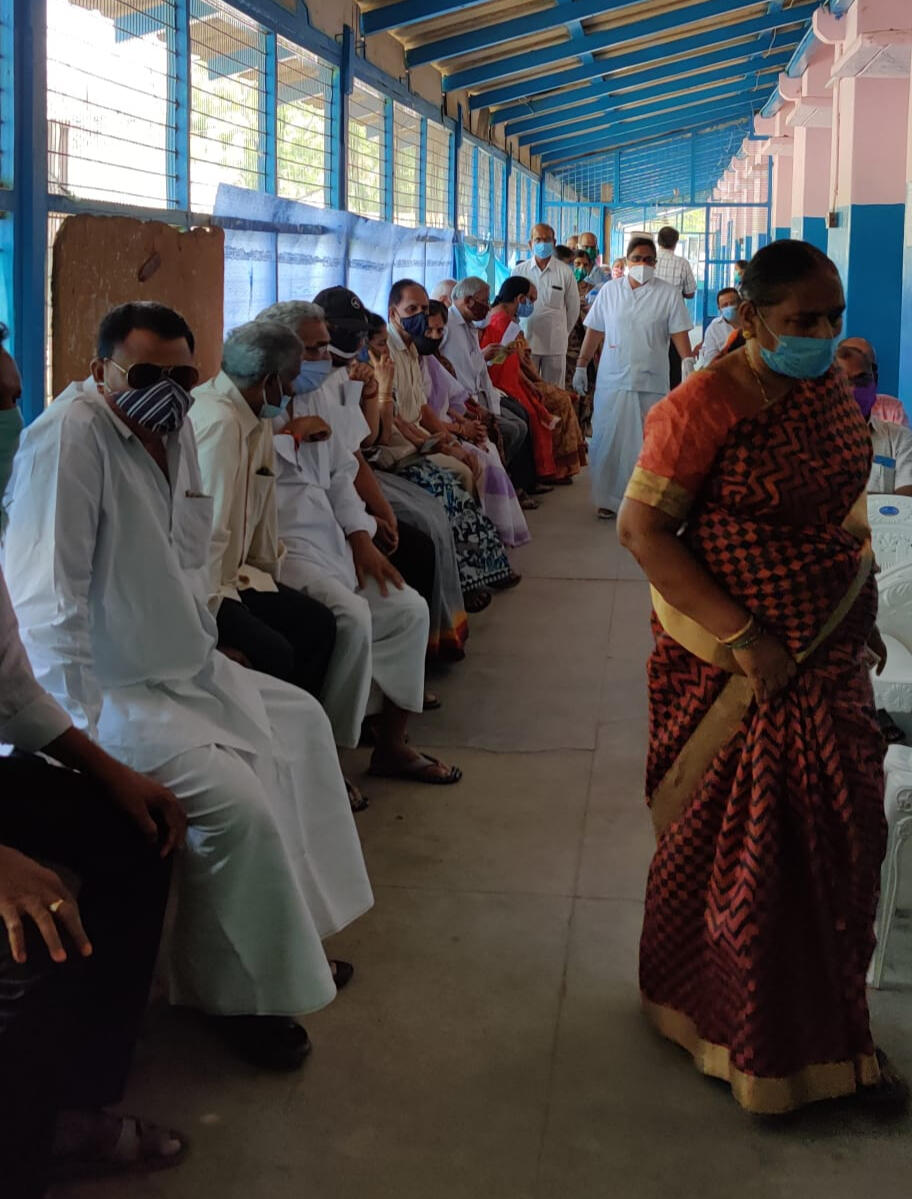 {Numerous people are sitting against a wall in a hospital in India. An older woman in a red saree is walking by.